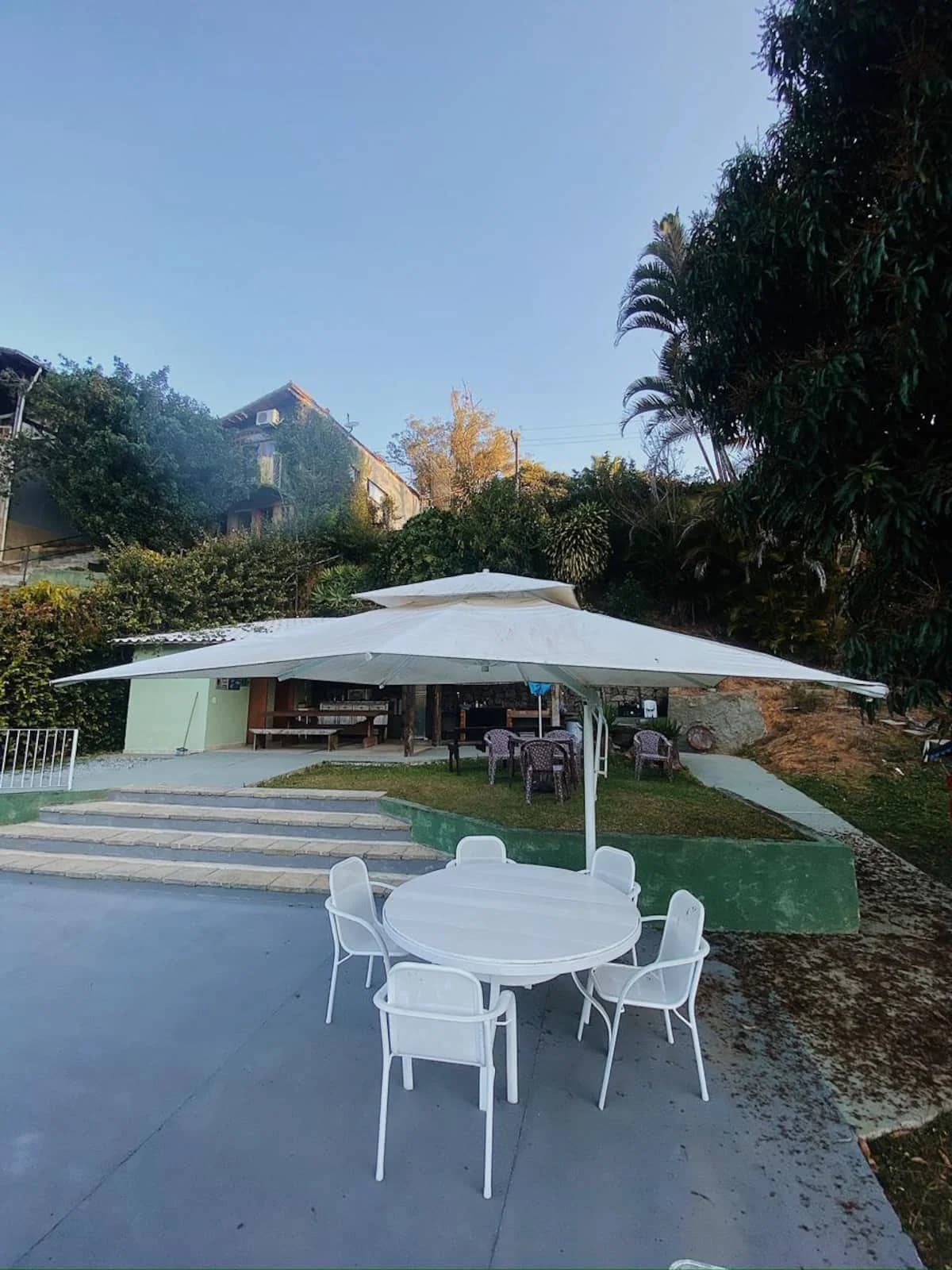 Área coberta com mesa redonda e guarda-sol, no piso de concreto junto ao gramado